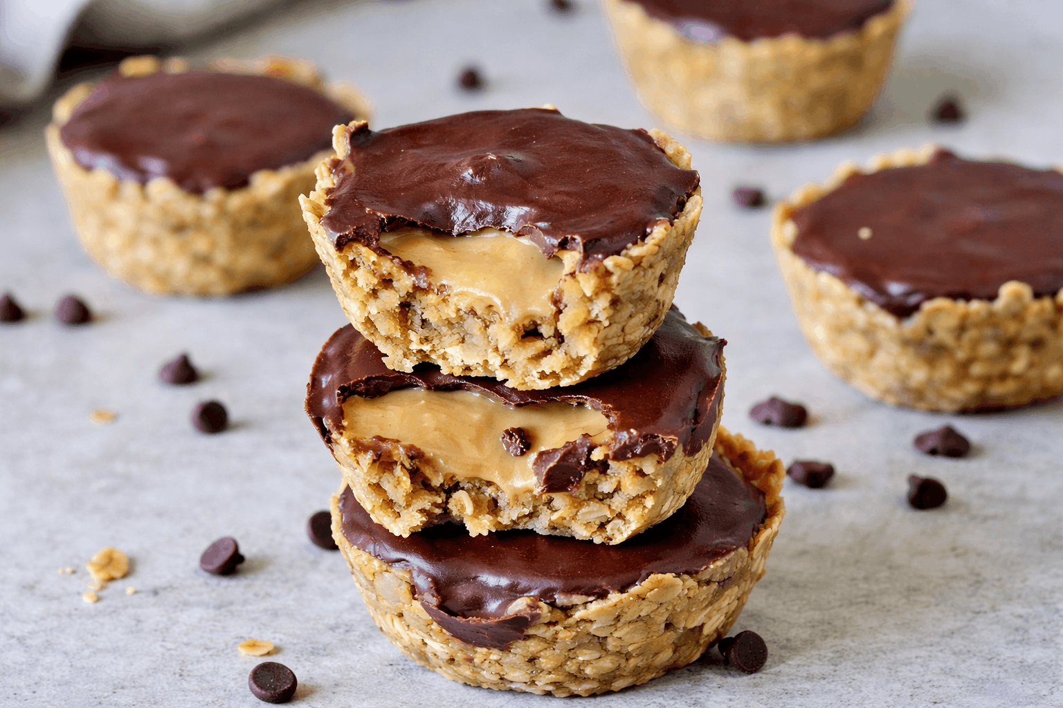Drei gestapelte Granola Cups ohne Zucker mit Mandelmus-Füllung und dunkler Schokoladenschicht auf hellem Steinuntergrund.